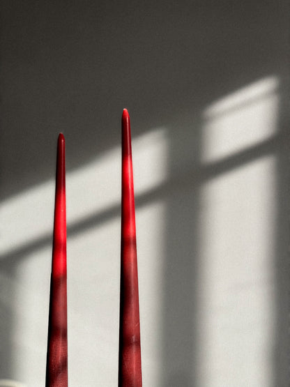 Two tall, slender red taper candles standing upright against a light gray wall, with soft window-light shadows cast across the background.