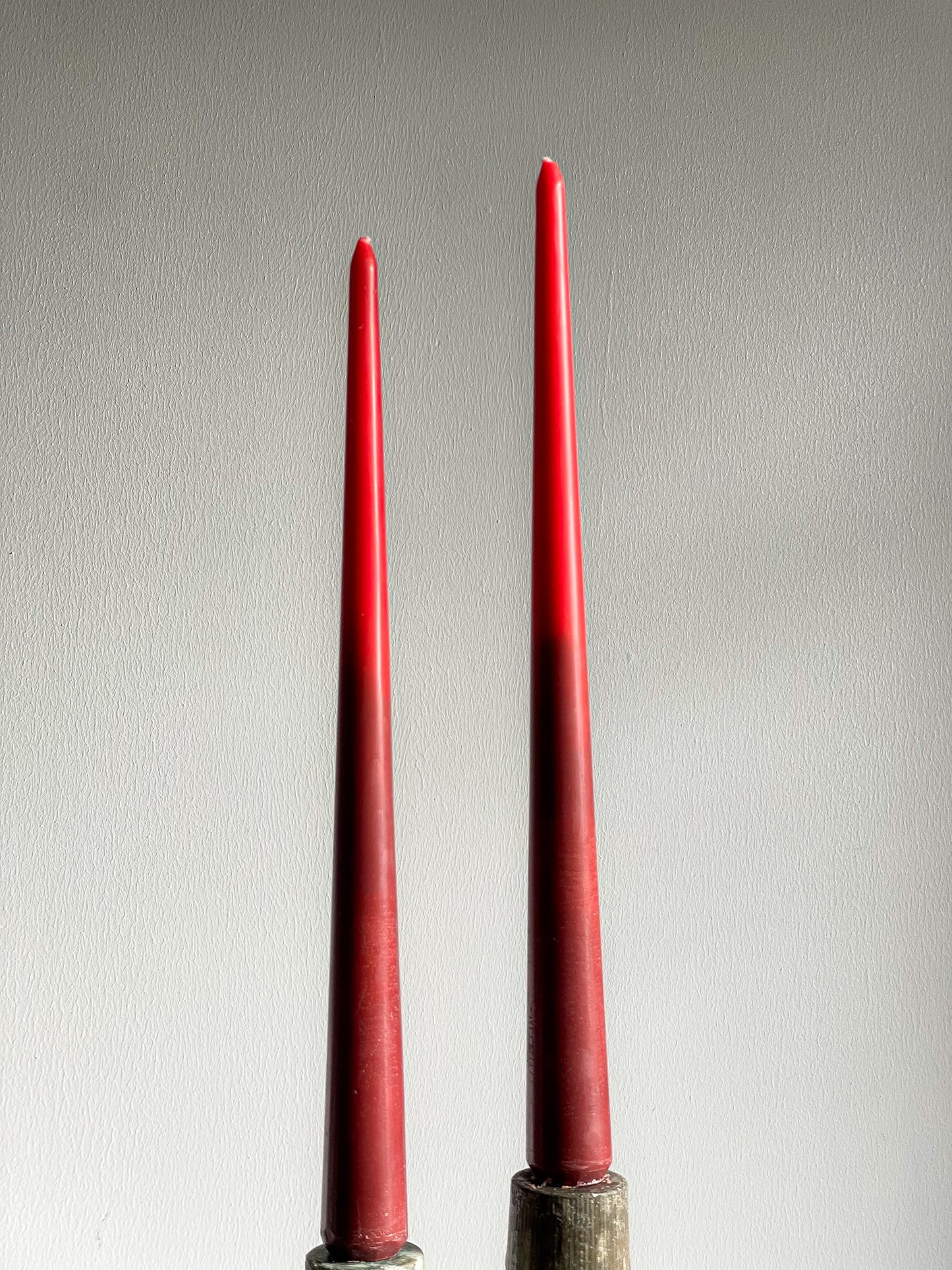 Two tall, slender red taper candles standing upright in dark metal holders against a plain light gray wall.