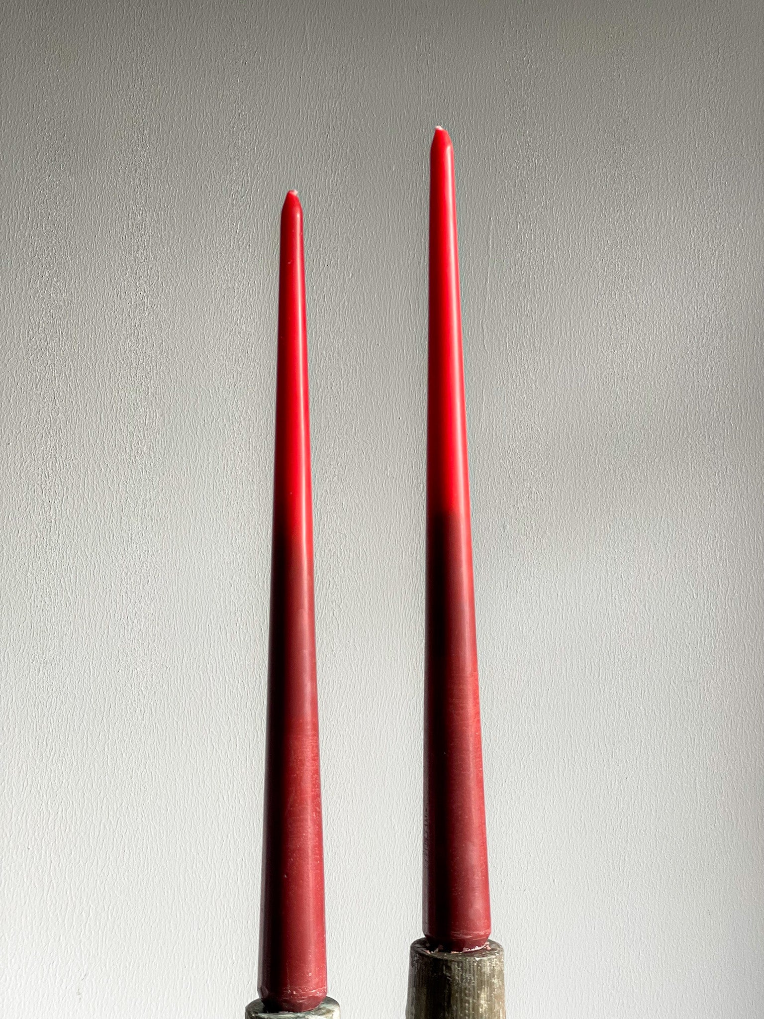 Two tall, slender red taper candles standing upright in dark metal holders against a plain light gray wall.