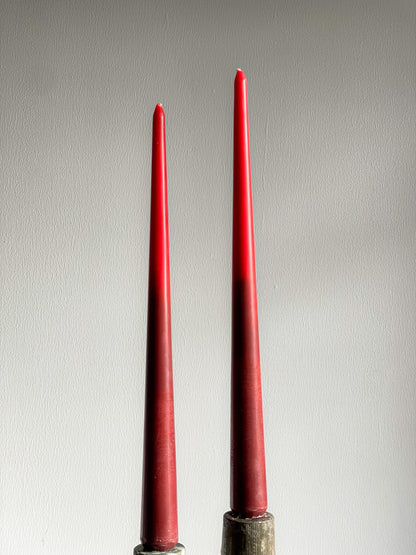 Two tall, slender red taper candles standing upright in dark metal holders against a plain light gray wall.