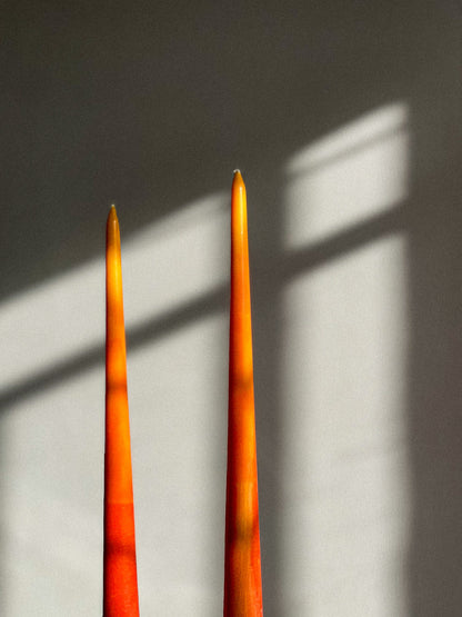 Two tall, slender orange taper candles standing upright against a gray wall with soft window-light shadows cast across it.
