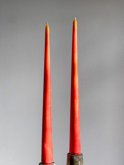 Two tall  orange-red taper candles standing upright in dark metal holders against a plain light gray wall.