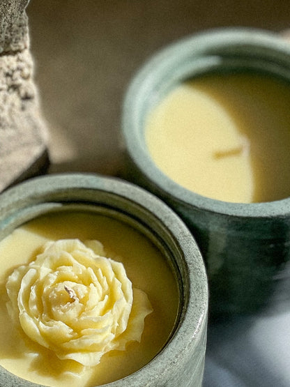 Two teal conccrete candle jars filled with pale yellow wax, one in sharp focus showing a sculpted peony-shaped wax bloom on top, and the other softly blurred in the background.